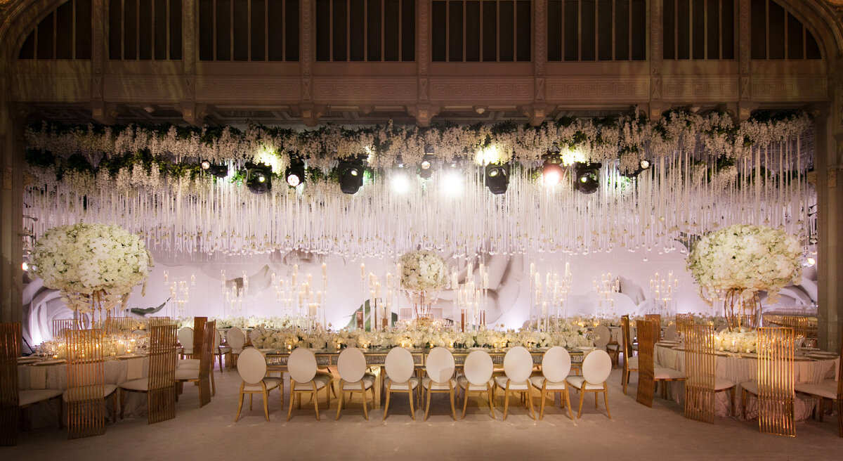 This stunning photo was captured at The New York Public Library in New York City by photographer Christian Oth of East West Films. The lavish indoor wedding reception showcases a long dining table with exquisite white chairs, set amidst a backdrop of abundant hanging white flowers and soft lighting. The romantic ambiance is further intensified by the tall floral centerpieces and flickering candles that adorn the space, adding a touch of elegance to the sophisticated decor.