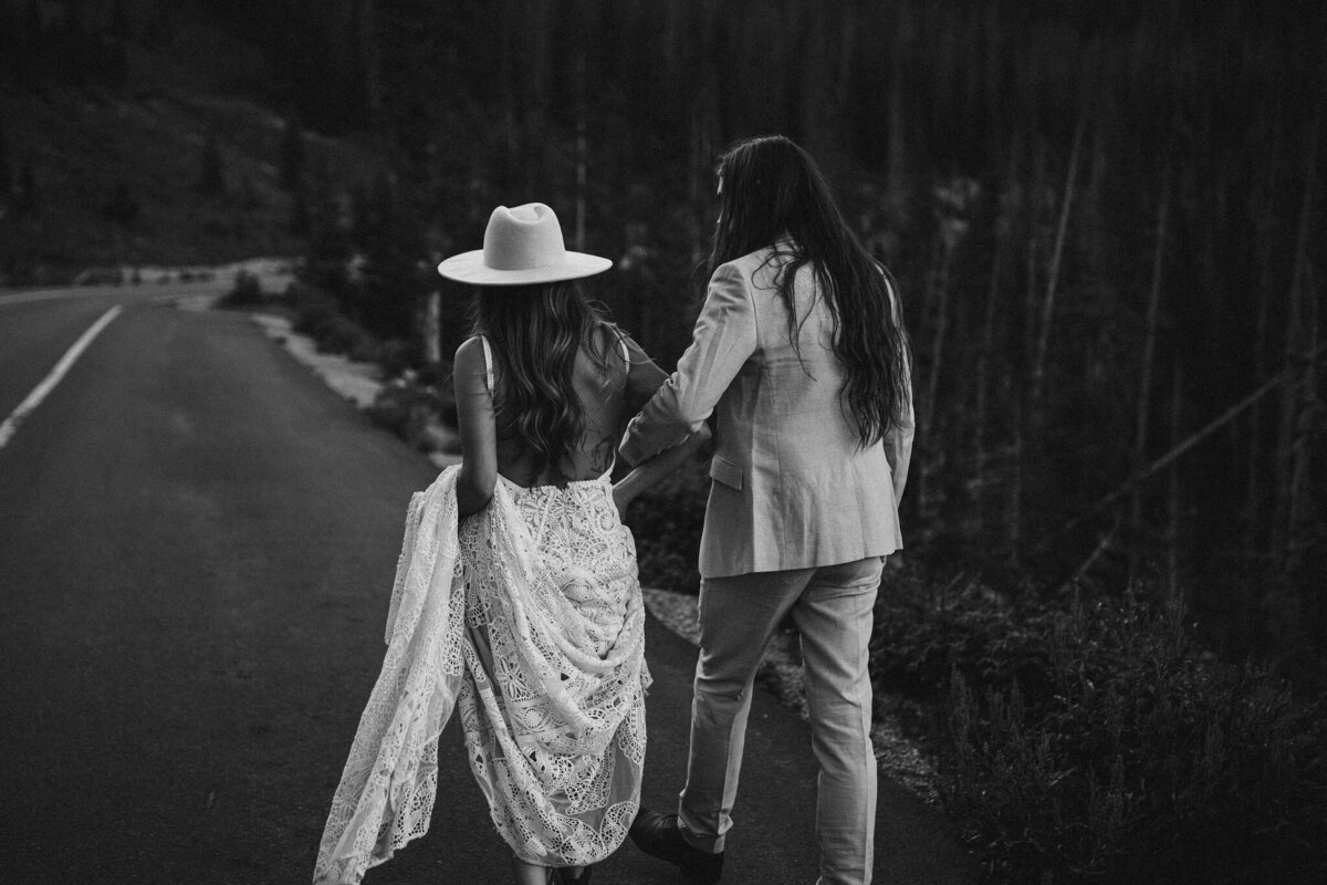 Black and white image of bride and groom wearing a suit and wedding gold walking along a mountain road.