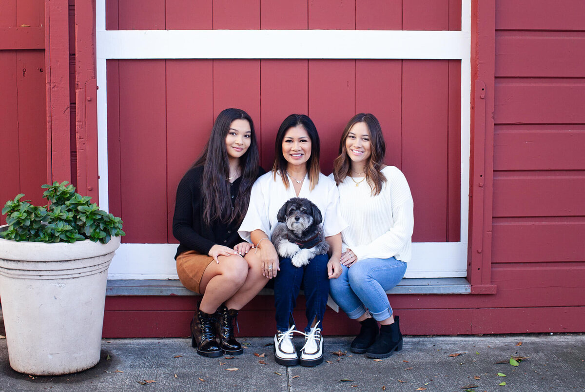 A mother with dark hair in a white shirt with her black dog sitting on her lap, sitting on a ledge beside her two daughters dressed in black and white, in front of a red and white barn door.