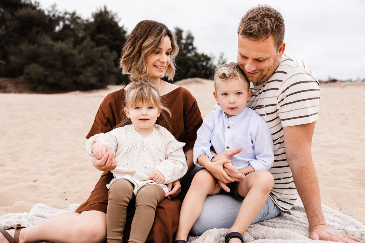 gezinsshoot mama in bruine jurk papa in gestreept shirt en zoon en dochter zitten op deken in het zand bij het eendenmeer in bergen