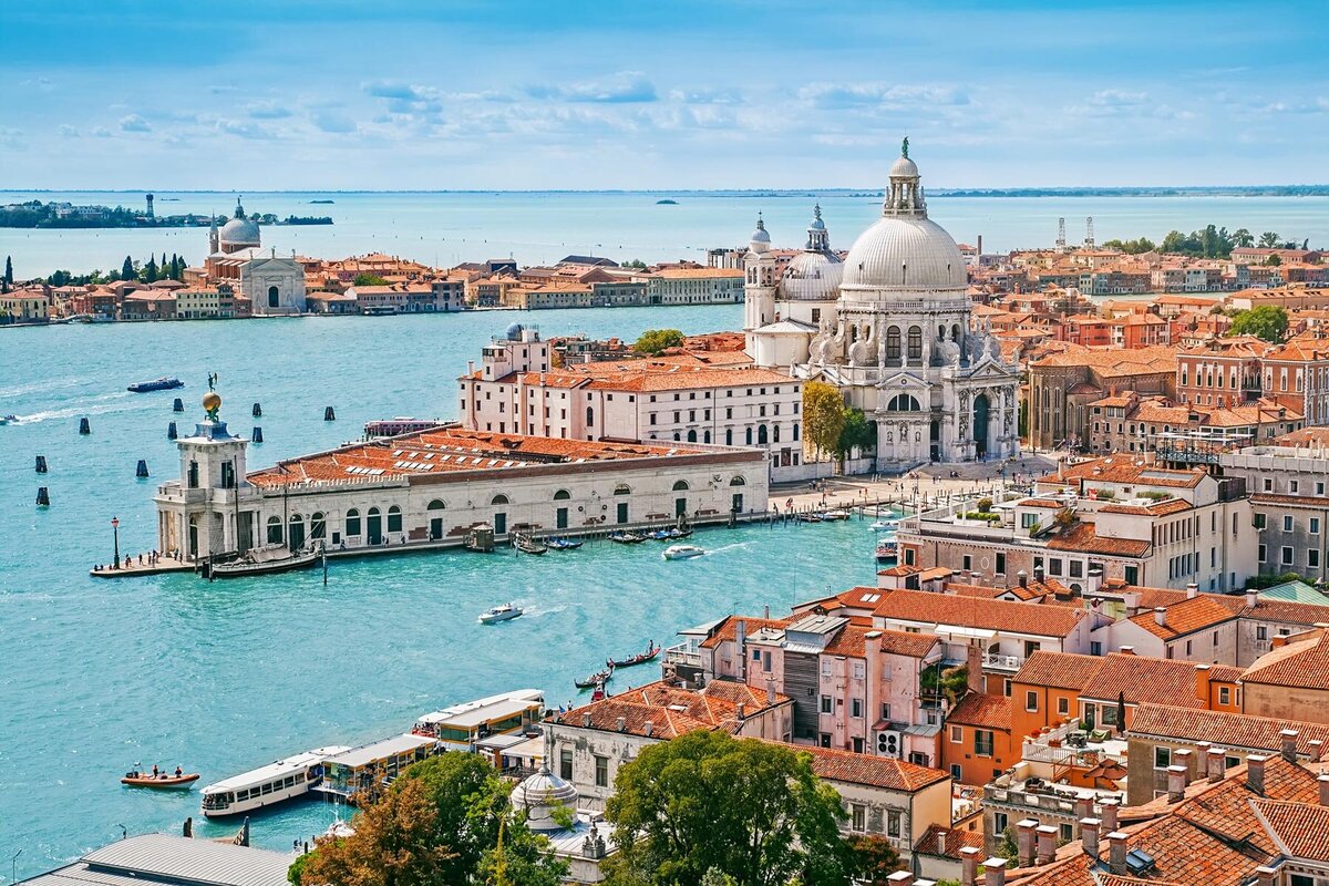 Aerial view of Venice, Italy, captured by a European travel specialist, showcasing the Grand Canal with boats, the prominent Basilica di Santa Maria della Salute, and surrounding historic buildings with terracotta roofs under a clear blue sky.