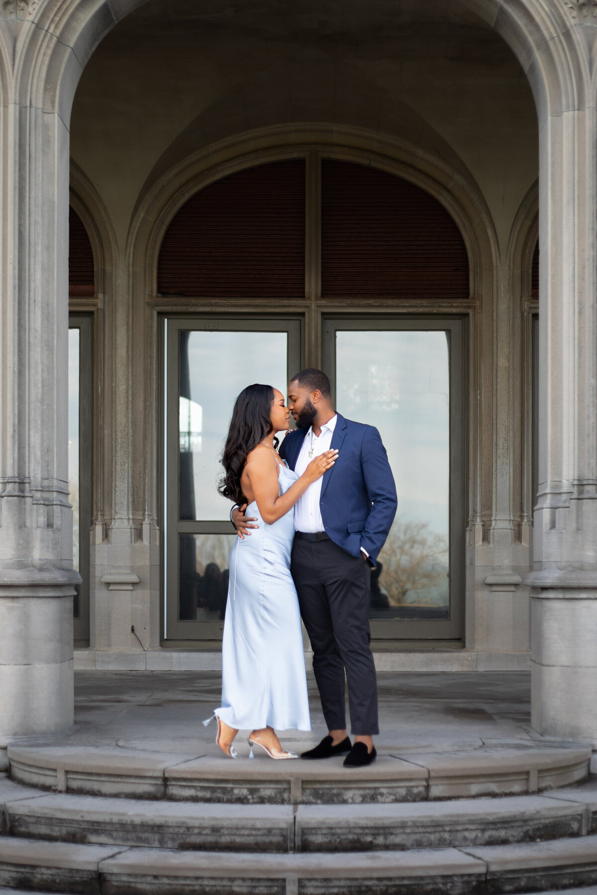 Engagement Photos Rhode Island | Kelsey Sheehan Photography Timeless Rhode Island Weddings | A couple shares a tender kiss in front of an elegant building with arched windows. The woman wears a light blue dress, and the man is in a navy suit. Romantic atmosphere.