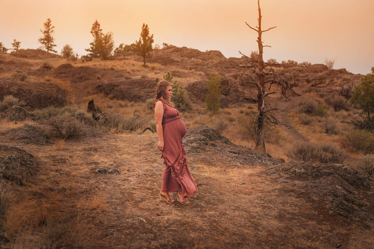 WIldfire season in Kamloops creates this stunning ambience for this Maternity photo. A woman wearing a rust coloured dress stands and looks off into the distance with a rocky terrain behind her.