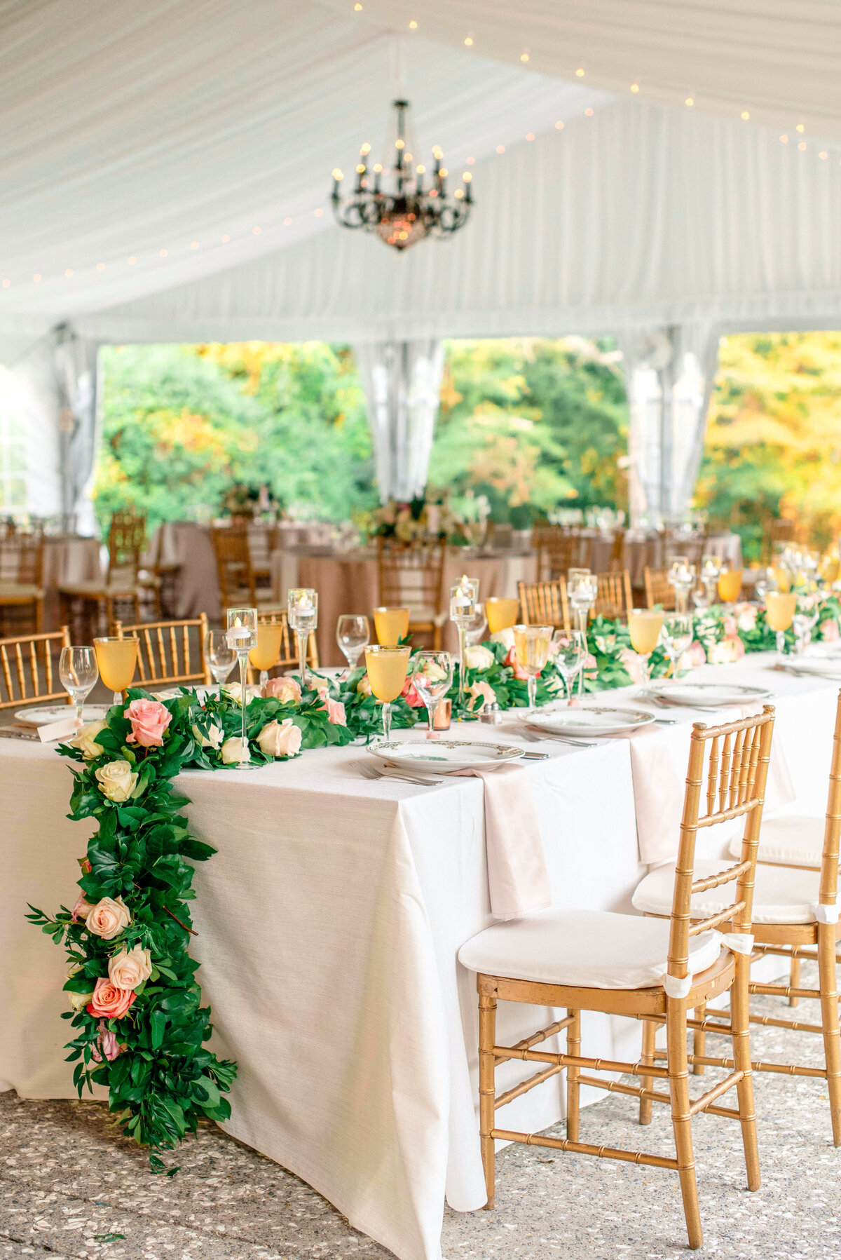 Elegant wedding reception setup under a white tent with gold chiavari chairs and tables adorned with garlands of greenery and pink roses. Champagne flutes and delicate tableware are arranged neatly.