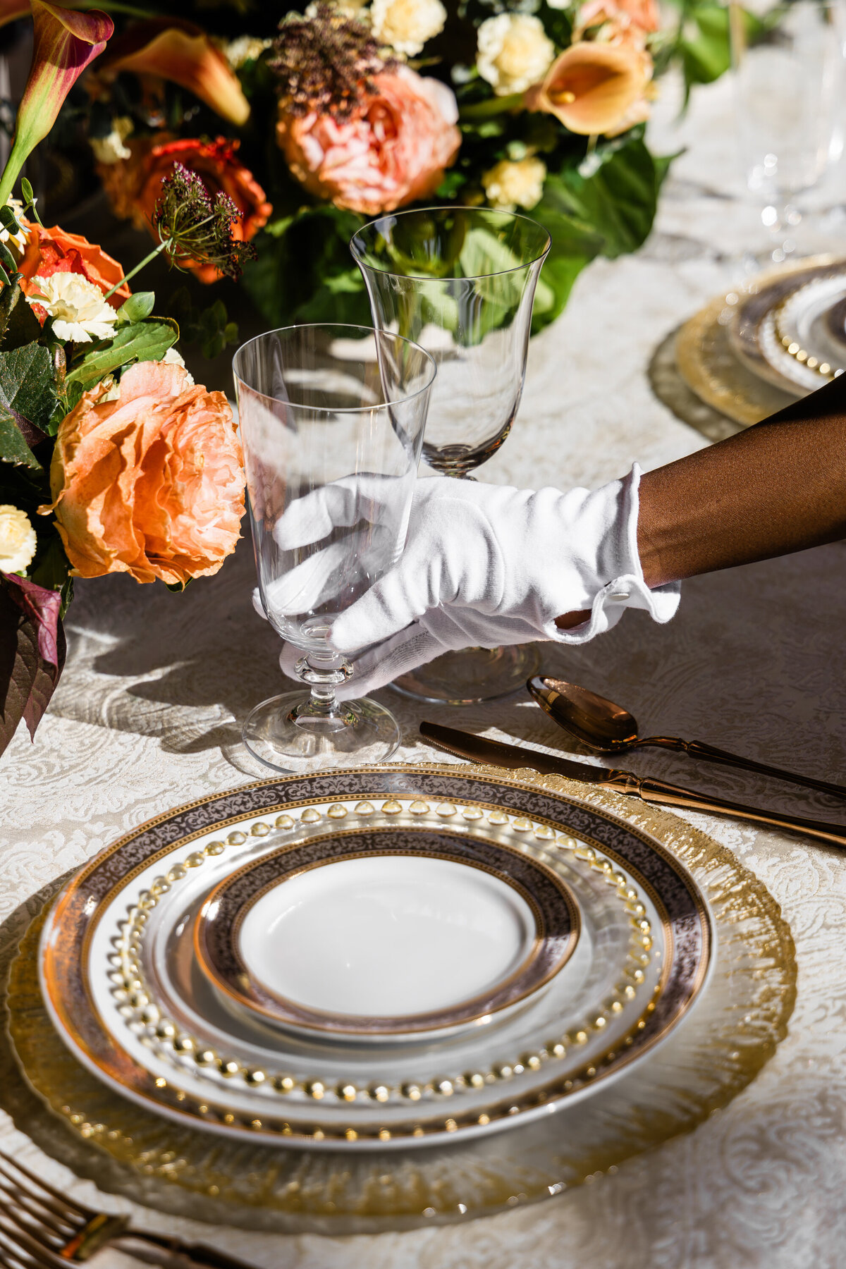 hand placing water glass next to  a four plate mixed metals place setting and a orange rose and calla lily centerpiece