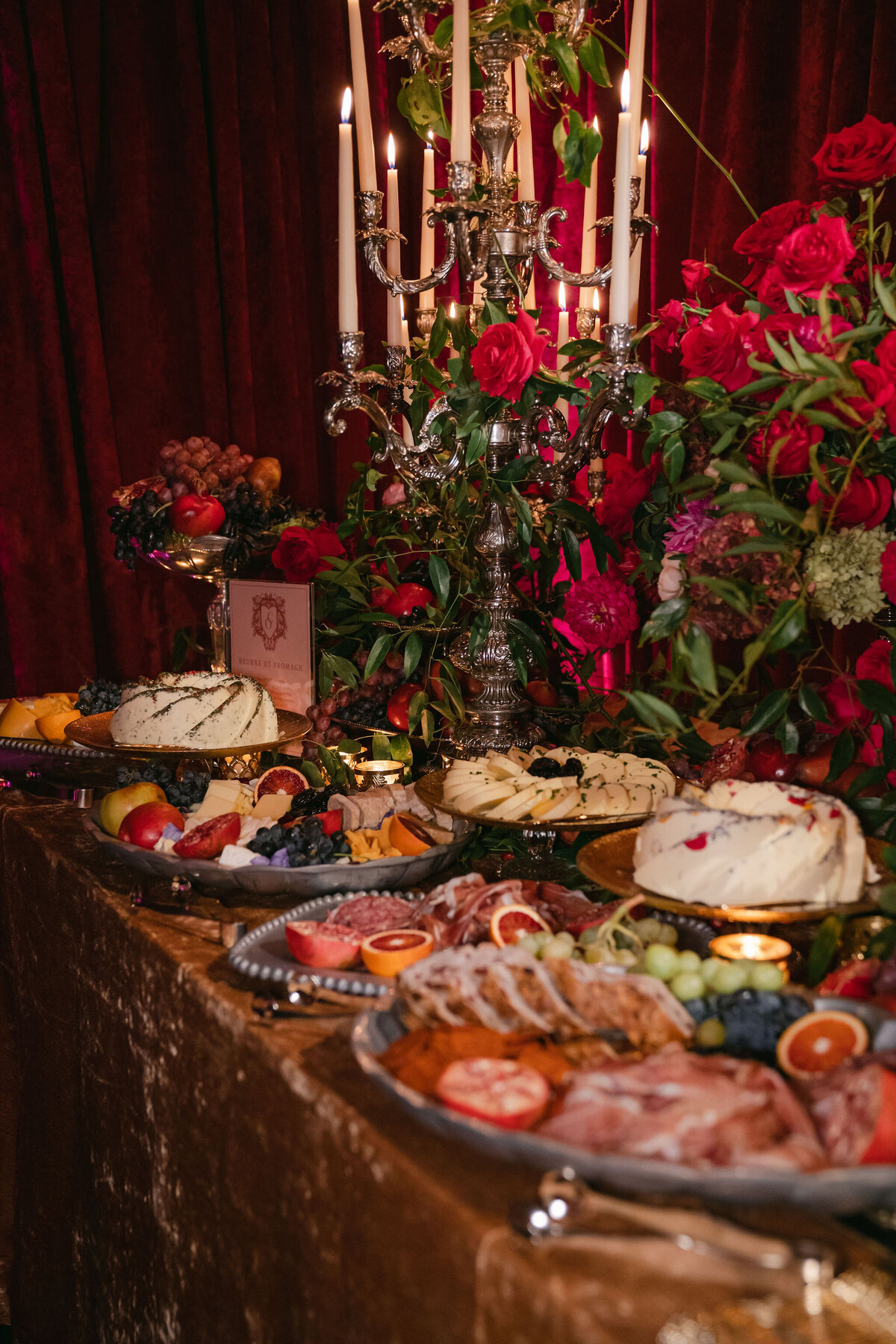A lavish spread of gourmet cheeses, fruits, and charcuterie arranged on a richly decorated table with a red tablecloth. The background features lush red roses and greenery, and a tall ornate candelabra with lit candles, creating an opulent ambiance.