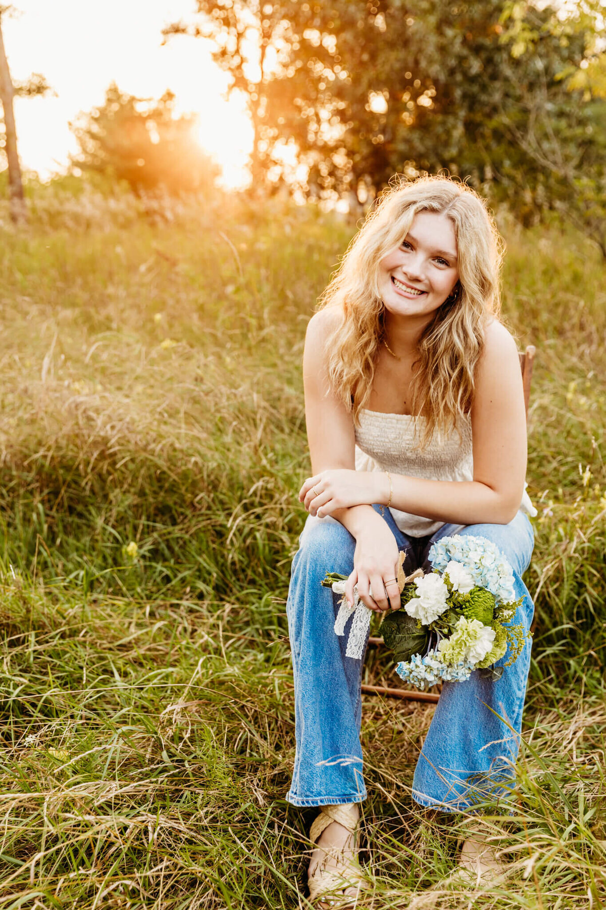 pretty blonde teenage girl in a white top and jeans sitting on a wooden chair in a field and holding a bouquet of flowers as the sun shines brightly behind her during her senior photo session