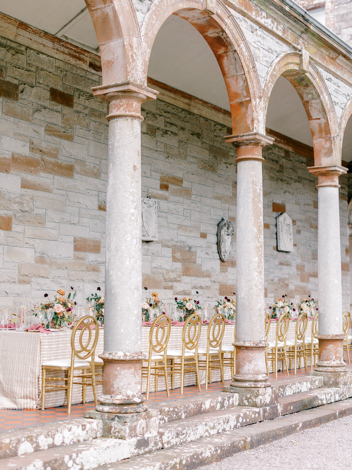 Photo of a white, pink, and maroon wedding reception table underneath stone archway photographed on an angle