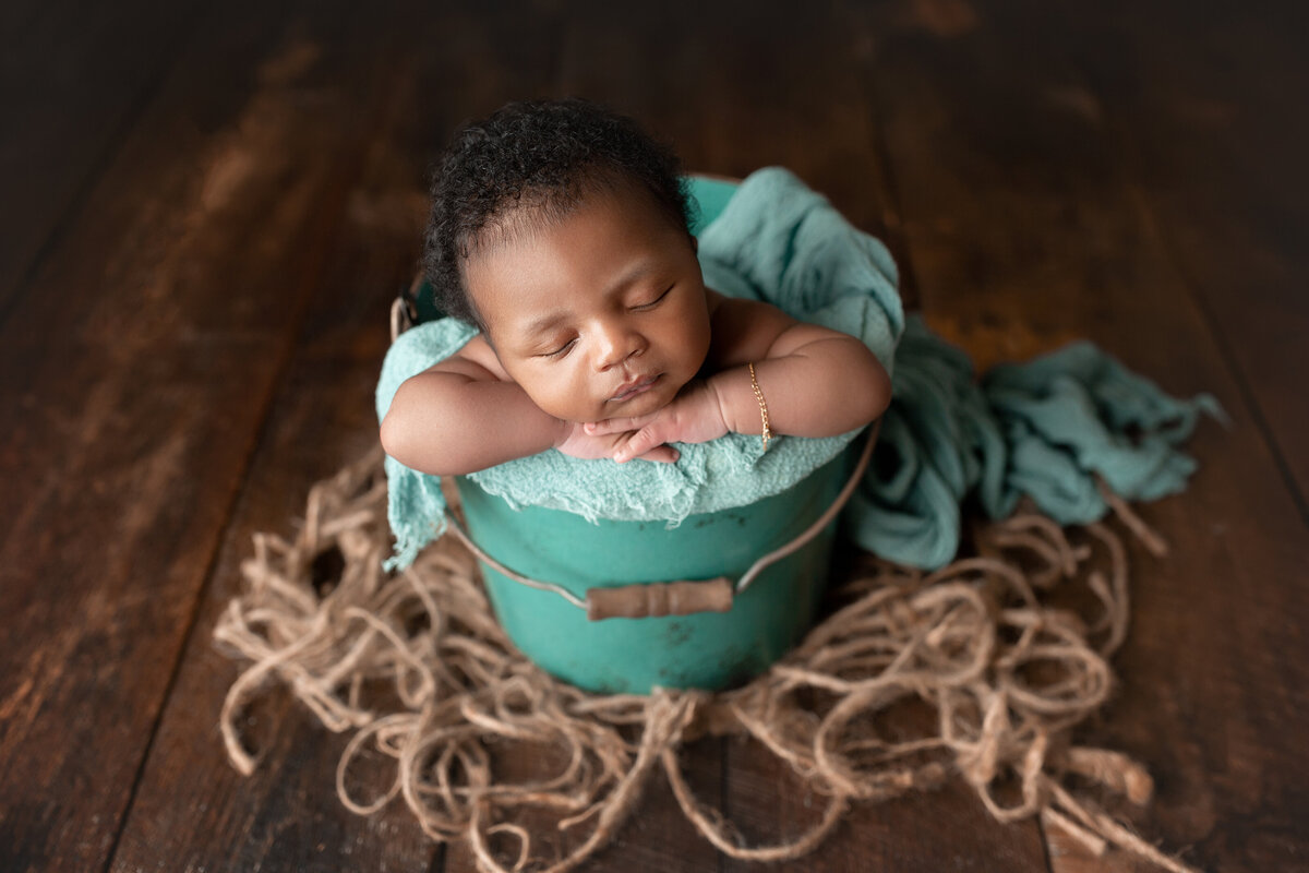 Black baby boy is sleeping in a teal bucket with matching teal blankets. Baby's hands are folded under his chin.