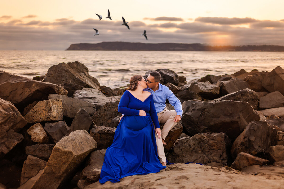 Maternity photographer, a couple sit on the rocks at the beach while the woman caresses her belly