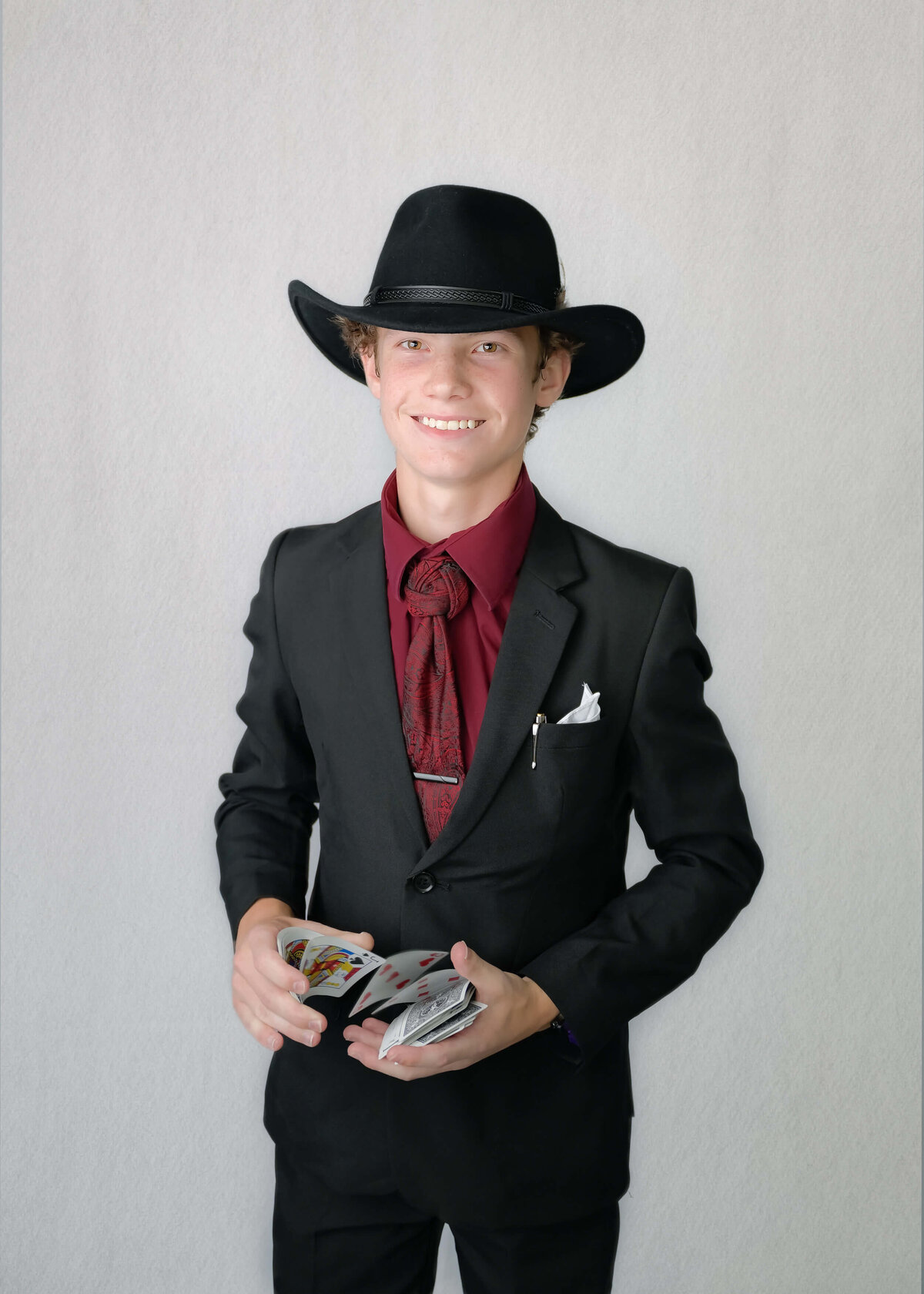 A studio-like family photo session of a teen dressed in a black suit, cowboy hat, and has a deck of cards ready to play Texas Hold 'Em. Family photo session in McKinney, TX by E. Benecki Photography.