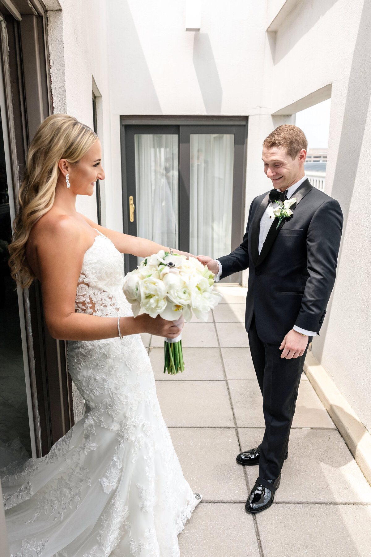A bride in a lace gown holds a bouquet of white flowers, standing near an open door on a terrace. She faces a groom in a black tuxedo who is smiling at her. They are outside, surrounded by tall building walls.