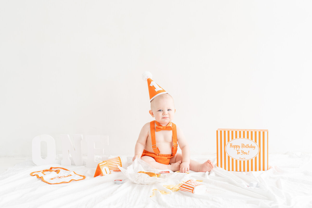 Little boy Whataburger themed first birthday session with decorations around him and eating burger and fries