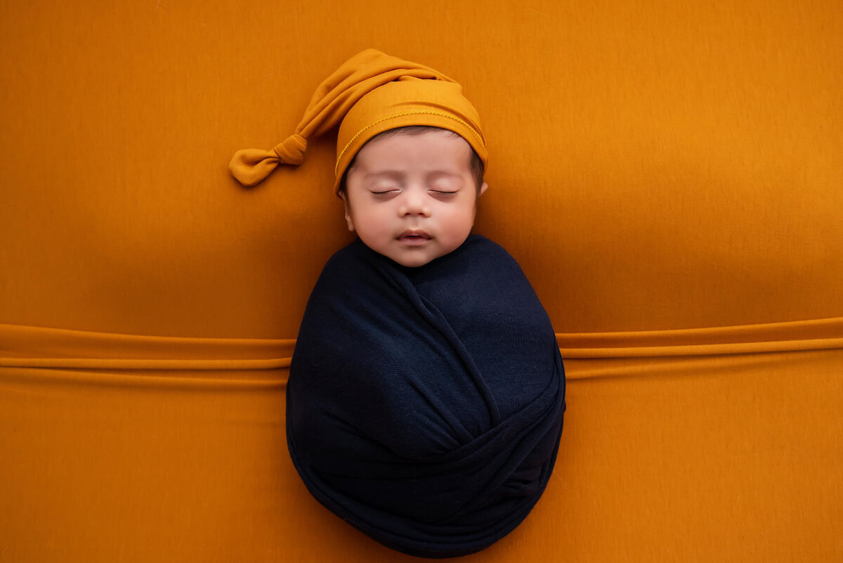 newborn boy wearing an orange sleepy hat on an orange background wrapped in navy blue for his newborn photography session in Burlington, Ontario