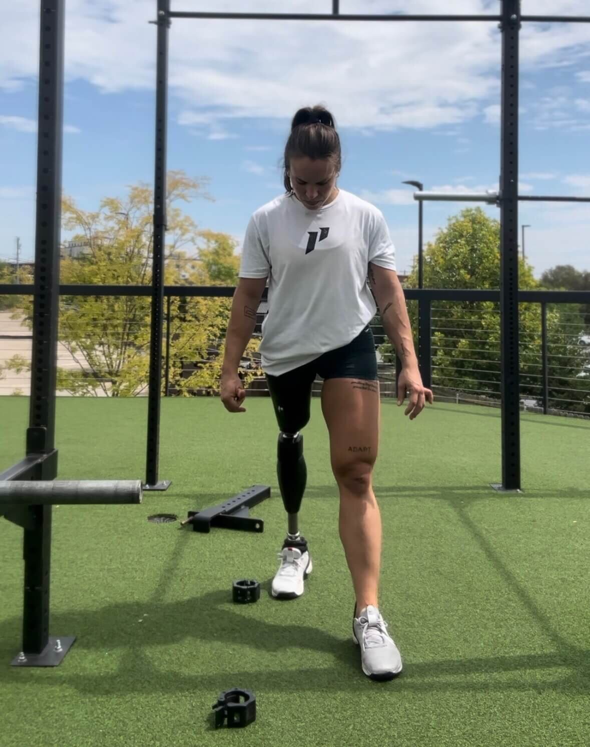 An athlete in a white performance t-shirt and black shorts trains at an outdoor gym facility with artificial turf. She is looking down while working out, wearing a prosthetic leg, with exercise equipment visible in the background including a rig and training accessories on the ground. The setting shows a sunny day with trees visible beyond the workout space.
