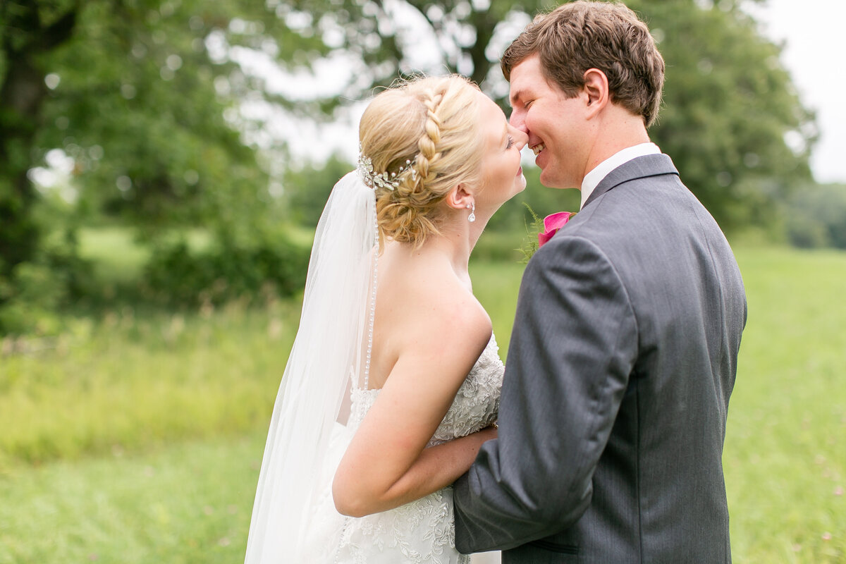 A white couple wearing wedding attire is standing in a field facing each other and moving in for a kiss.