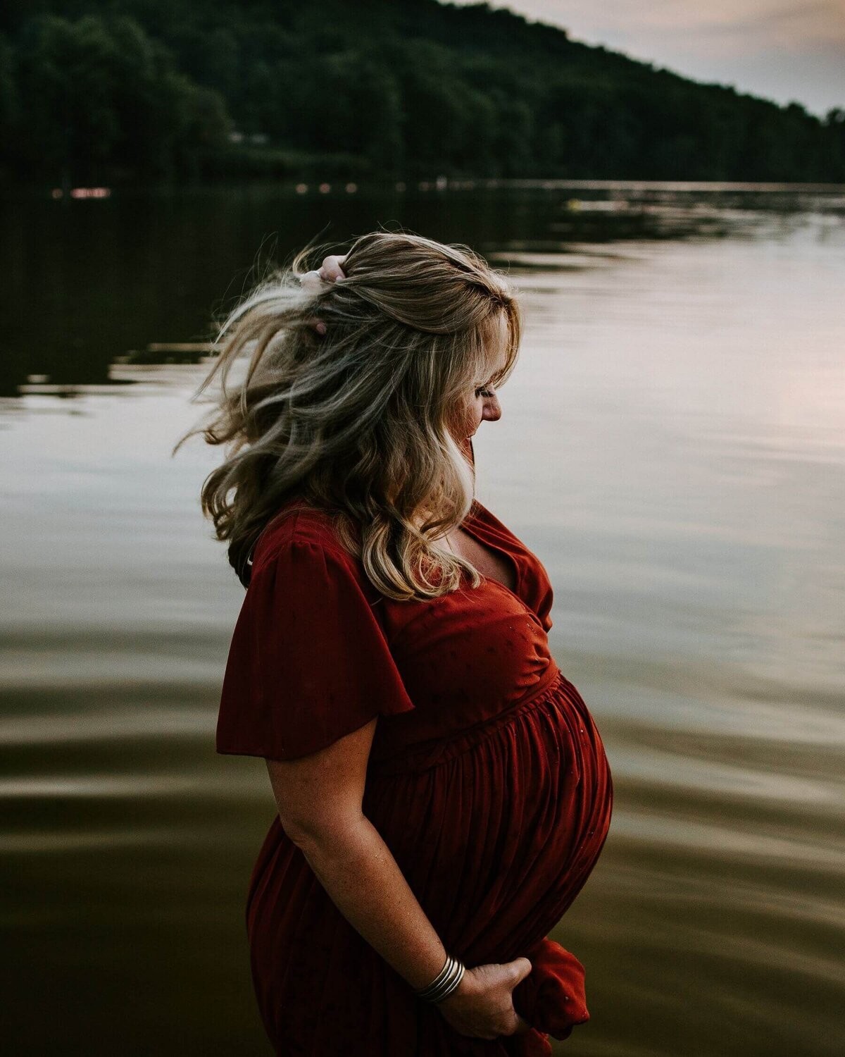A pregnant woman in a red dress poses elegantly by a lake, captured by a Pittsburgh maternity photographer.