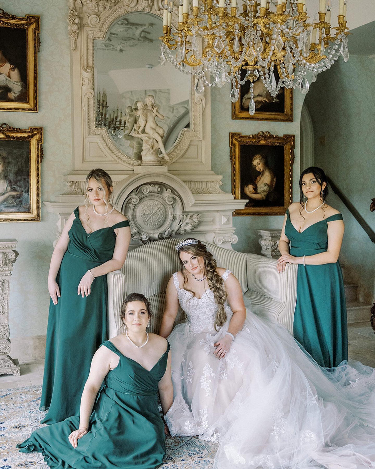 Bride wearing her bridal gown sitting down on a white chair while her girls wearing green gowns standing beside her