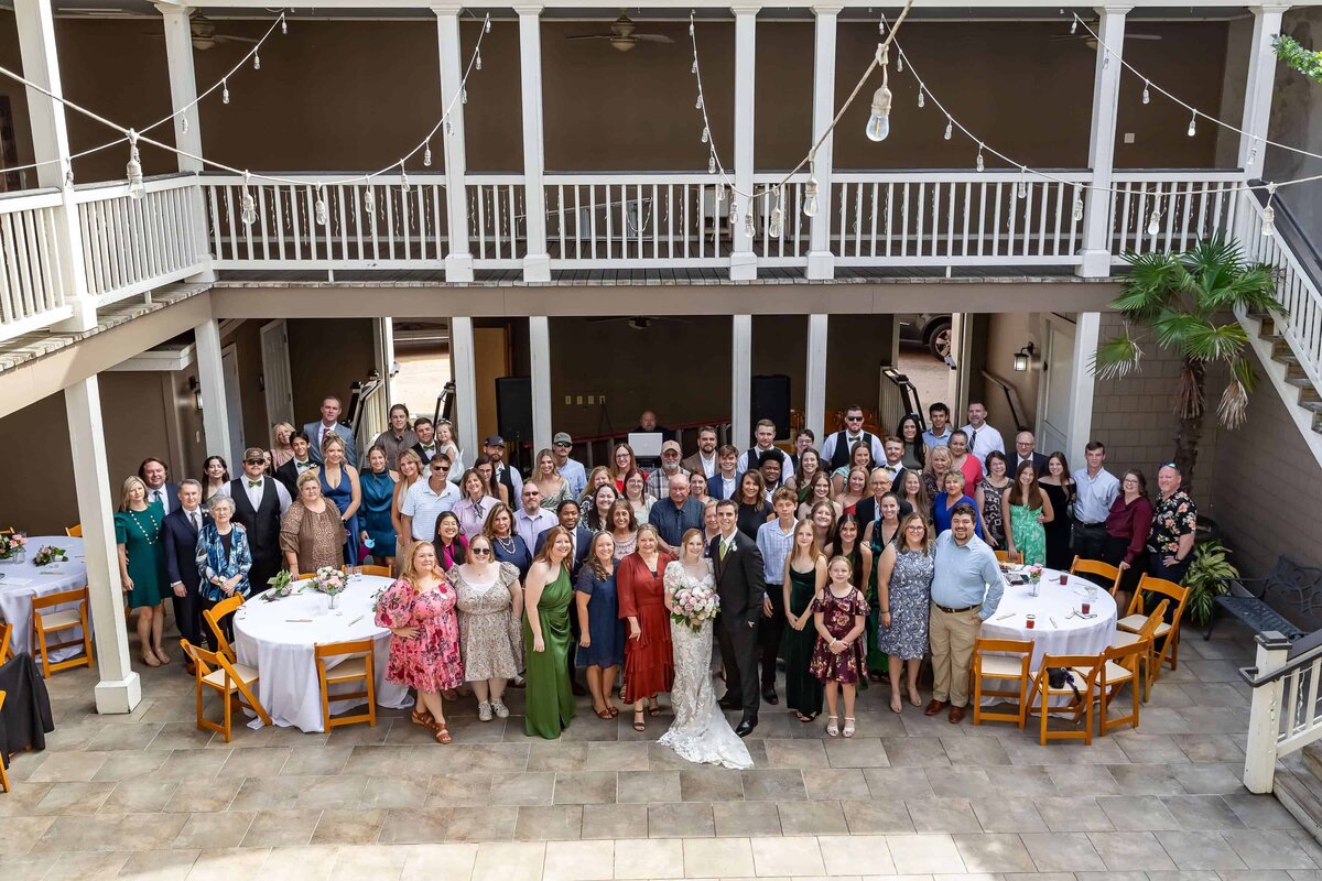 A large wedding group photo is taken in a courtyard with string lights and round tables. The bride and groom stand at the center, surrounded by guests smiling at the camera. The setting is festive and elegant.