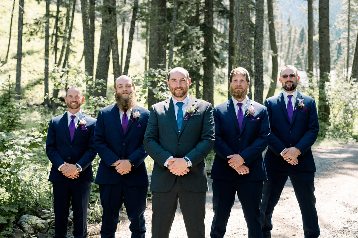 Groom and groomsmen in black tuxedos with violet ties, standing with hands clasped among towering pine trees during a summer wedding at The Woodlands at Cottonwood Canyon in Bozeman, MT.
