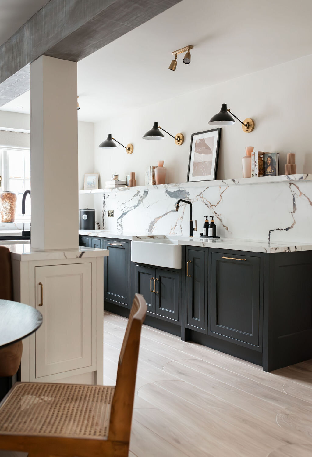 A modern kitchen featuring dark lower cabinets with a white marble countertop and backsplash. Wall-mounted black lights and neutral-toned decor add to the aesthetic. The kitchen has a farmhouse sink, sleek faucets, and light wood flooring.