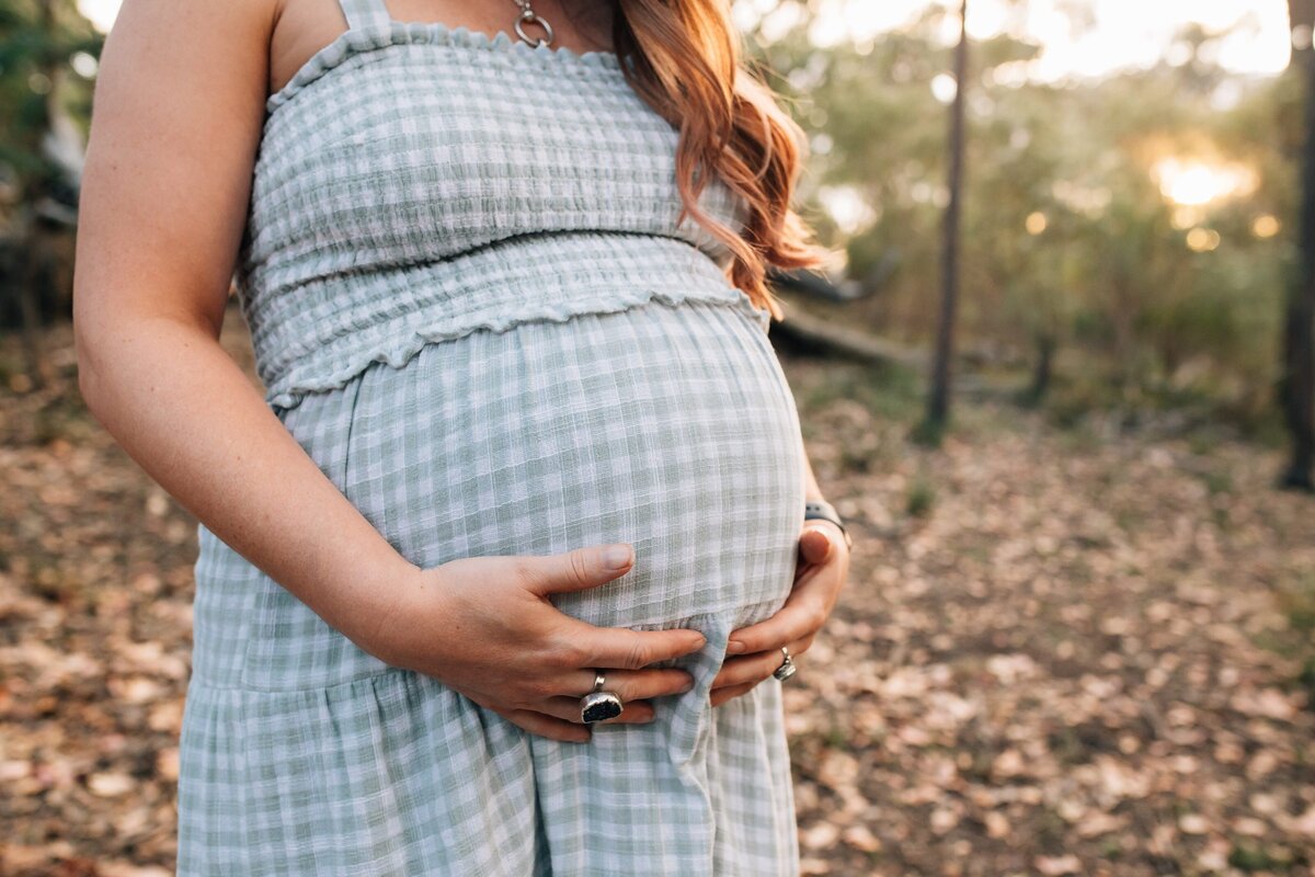 Pregnant woman holding belly, wearing blue checked dress outside during sunset.