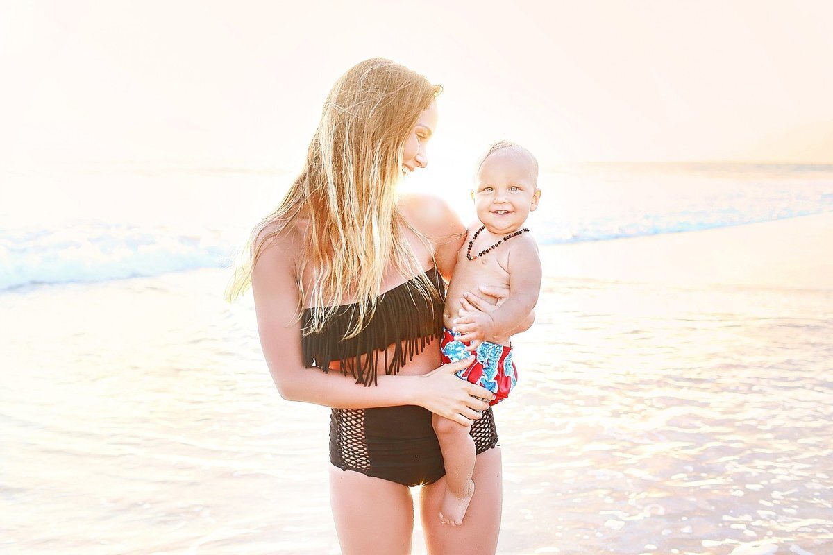 Young mother with black boho bikini holds her toddler on her hip and glances down at him, smiling on the beach at sunset