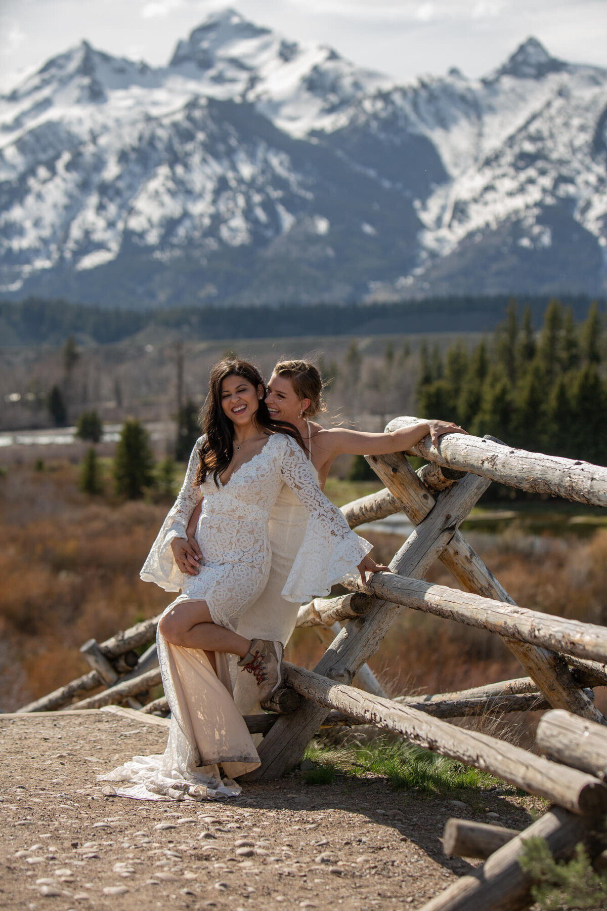 Two brides stand leaning against a wooden fence smiling at their Wyoming Elopement photographer