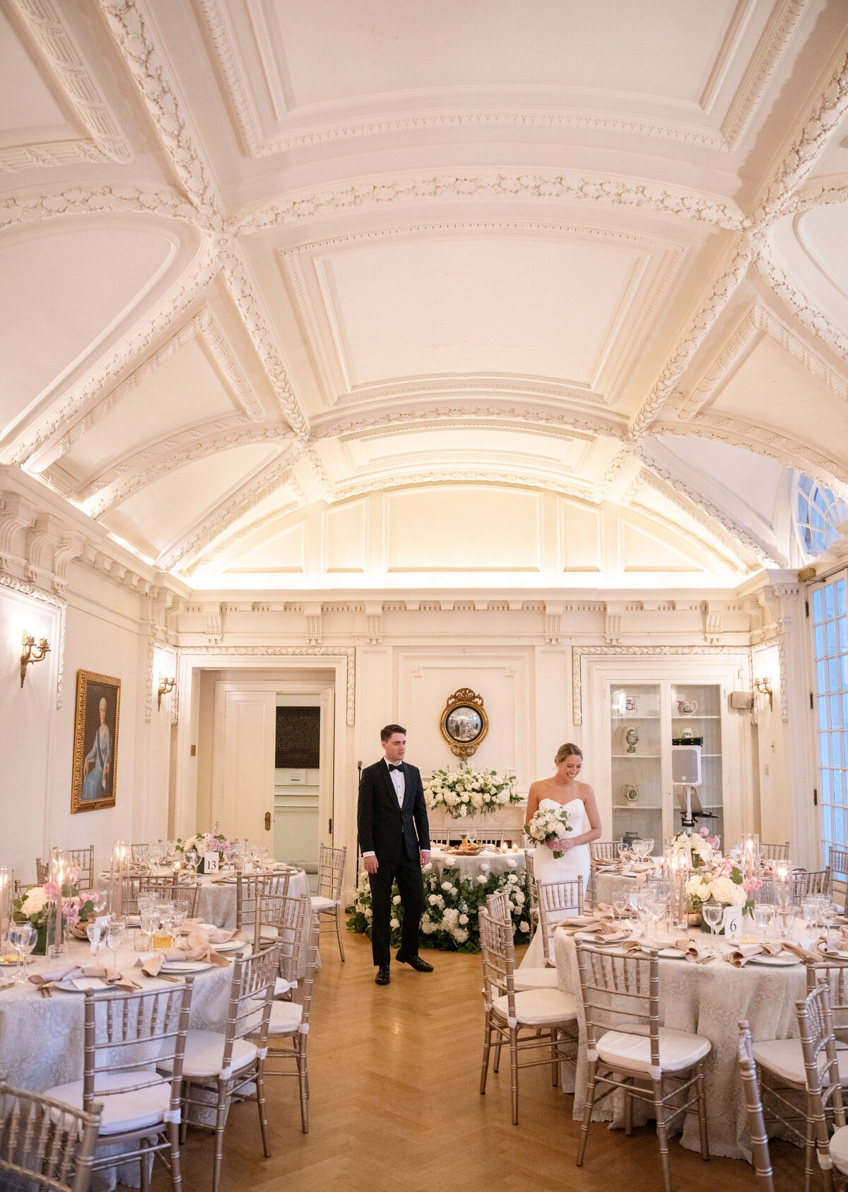 A couple in formal attire stands in an elegantly decorated, bright reception room. The room features ornate white ceilings, floral arrangements, round tables with white tablecloths, and chairs. Candles and table settings are visible.