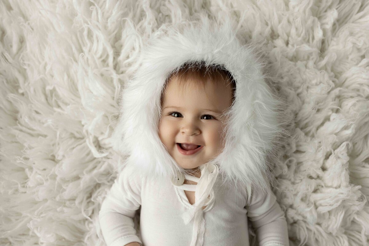 Baby girl in white onesie and faux-fur trimmed bonnet laying on fuzzy rug and smiling at the camera. Aerial image.