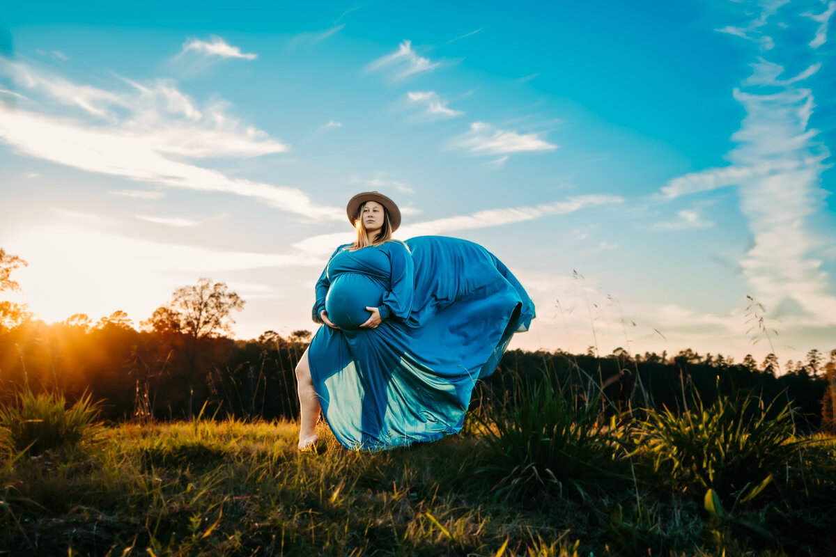Pregnant mother with dress blowing in the wind on a hill top at sunset wearing a hat