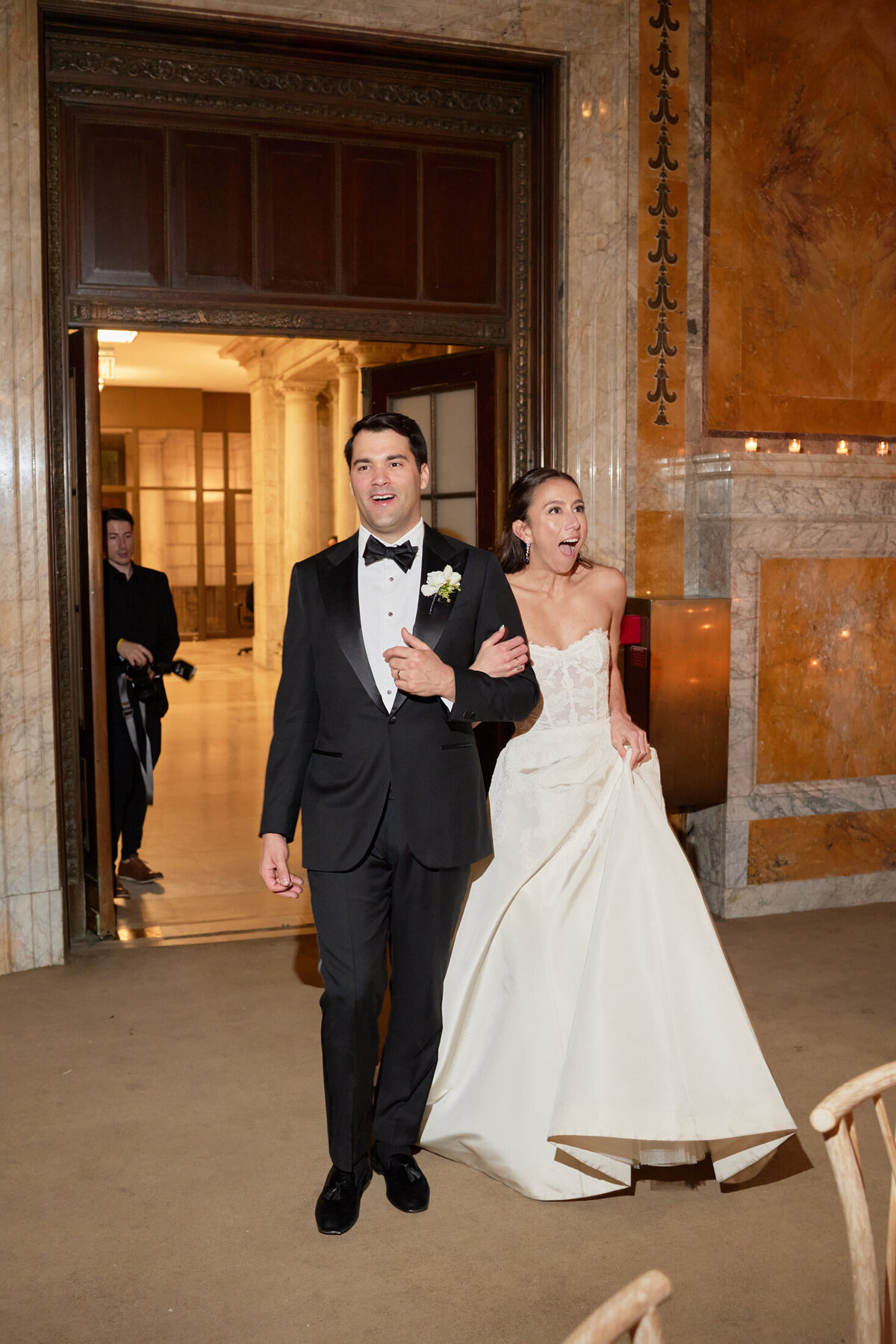 The photo was taken at the iconic New York Public Library in New York City by photographer Shawn Connell. A couple dressed in formal wedding attire, with the groom in a black tuxedo and the bride in a strapless white wedding dress, enters a lavishly decorated room within the library. Both of them look joyful, with the bride exuding excitement as they take in the ornate walls and stunning lighting of the room. Videography by Aaron Navak Films.