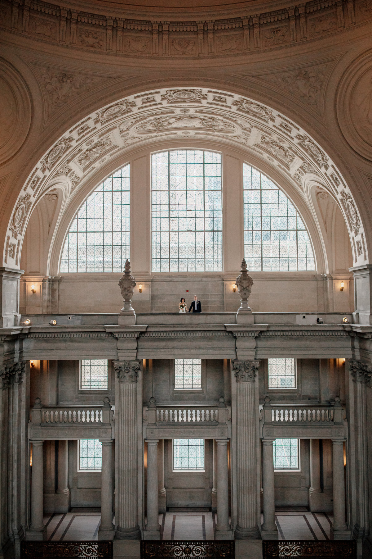 Couple holds hands in semi circle window archway. Wide angle wedding photo  of San Francisco City Hall