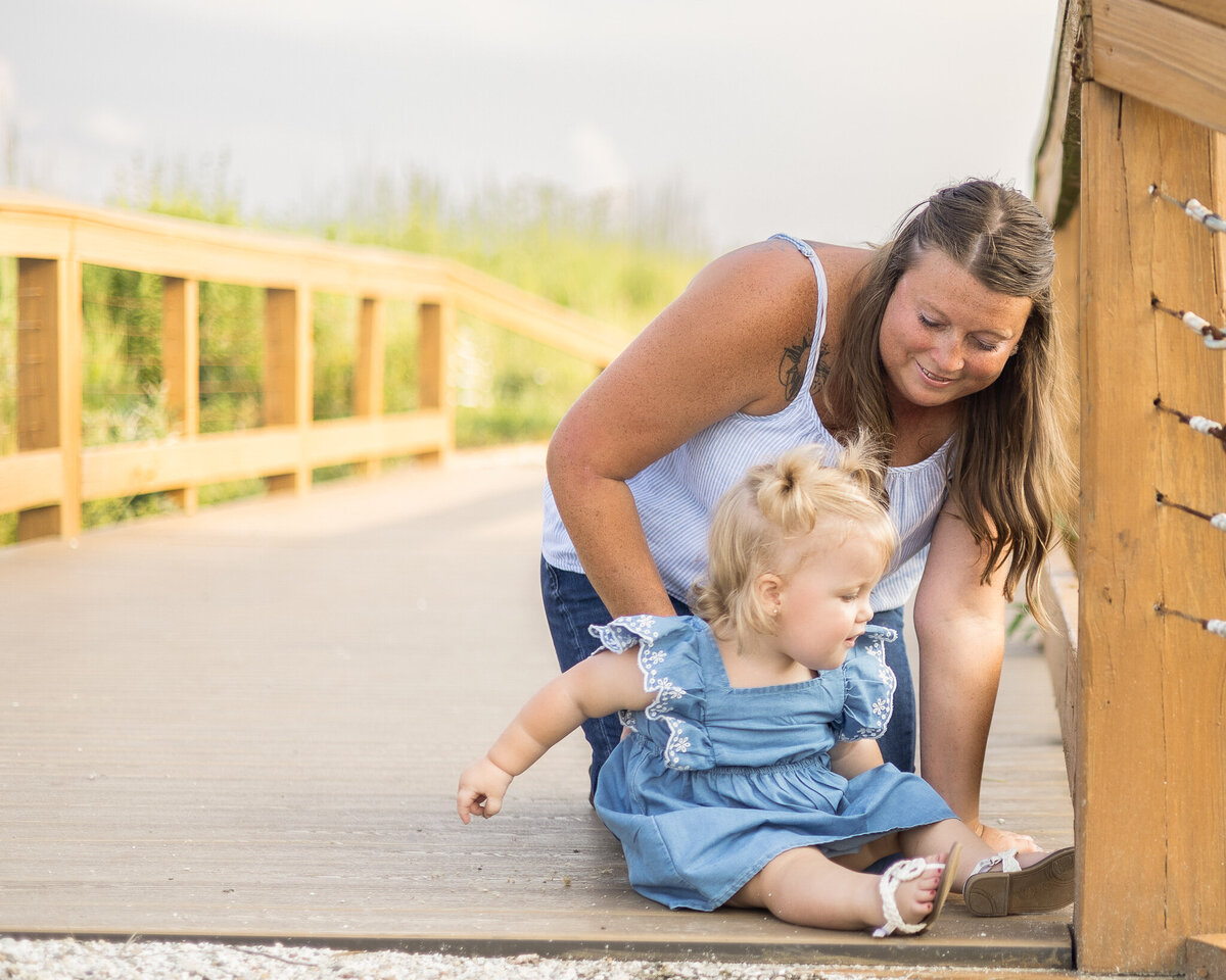 Mom and daughter sitting on a bridge at a wildlife sanctuary.  Mom is a brunette, wearing a white tank top, blue jeans, smiling down at her daughter.  Daughter has blond hair wearing a blue dress and sandals, smiling at the camera