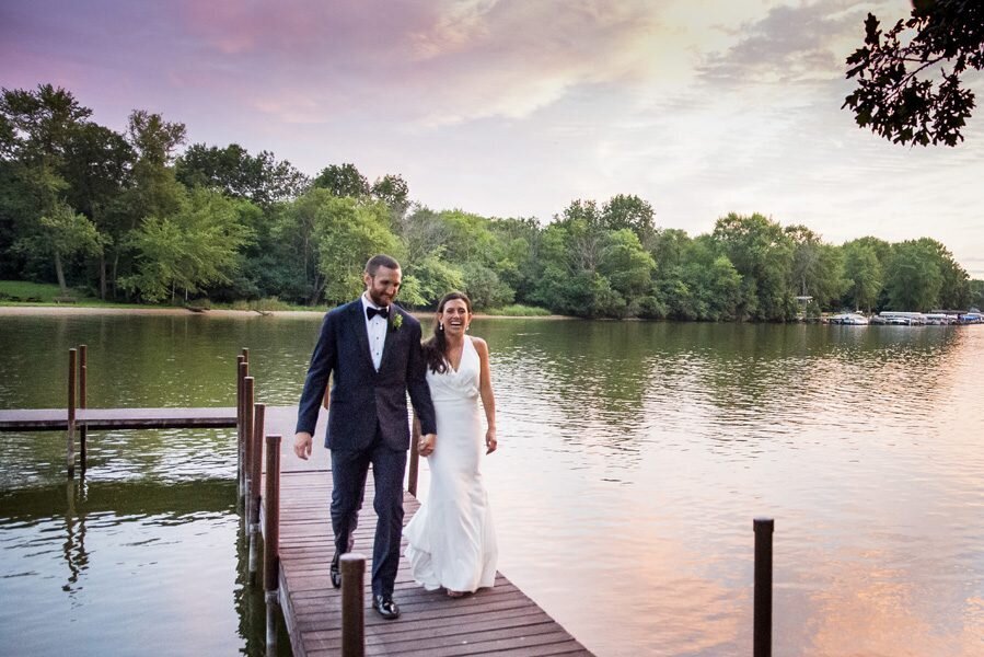 A bride and groom walk hand-in-hand toward the camera on the dock of a lake.