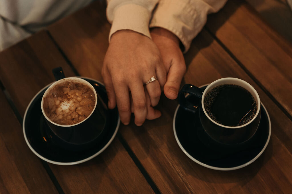 Overhead photo of couple holding hands, showing off engagement ring with coffee cups on either side.
