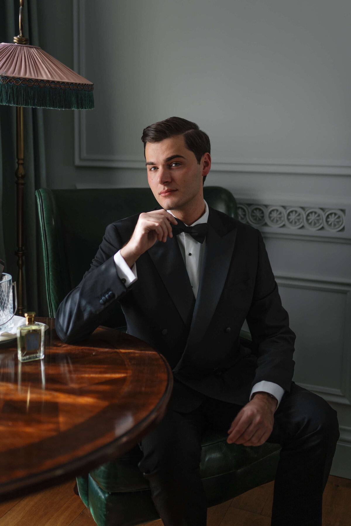 groom getting ready for his chic london wedding at the ned hotel sitting in his bedroom at a table in his suit and bow tie