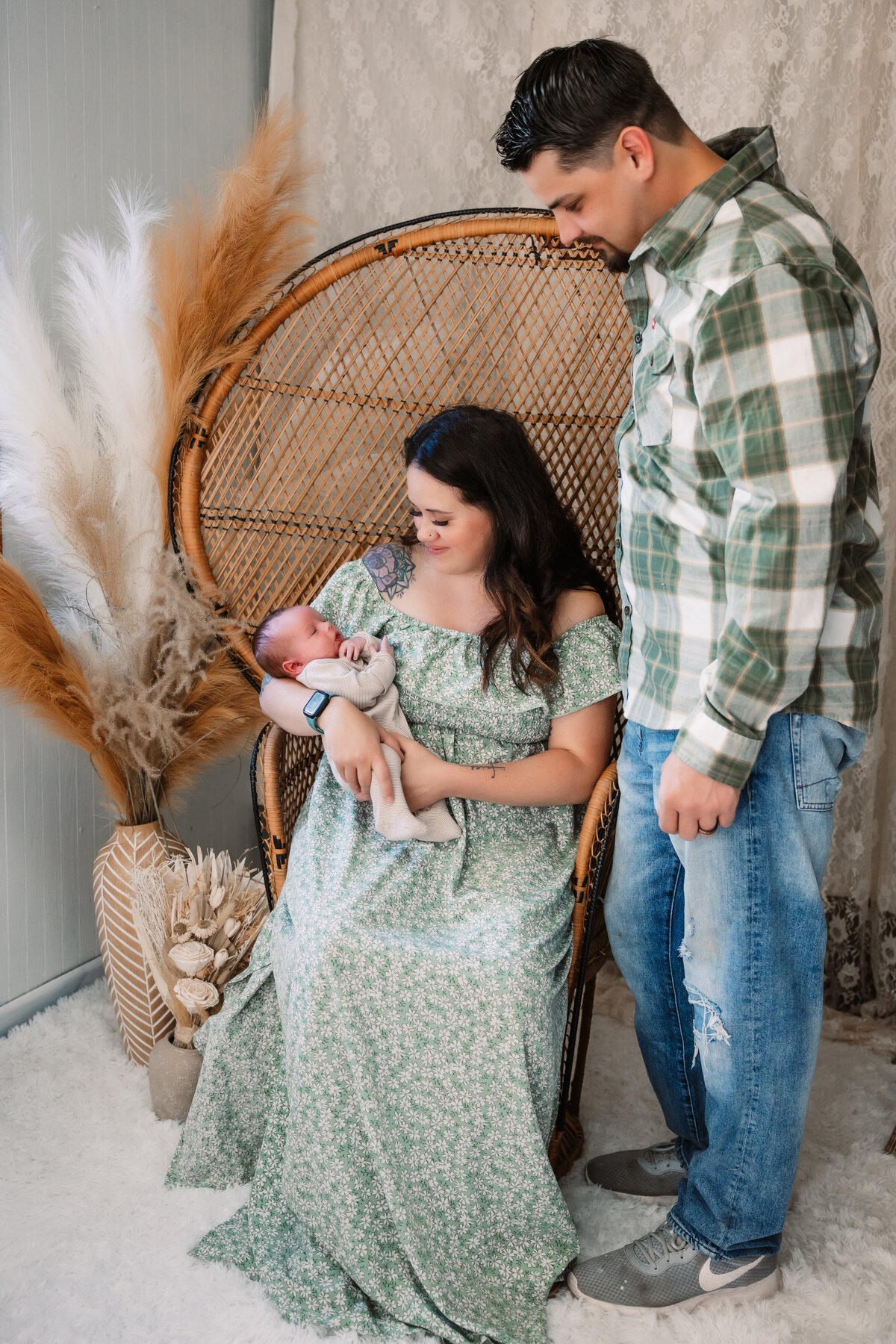 A father stands over his wife sitting in a wicker chair holding their newborn baby