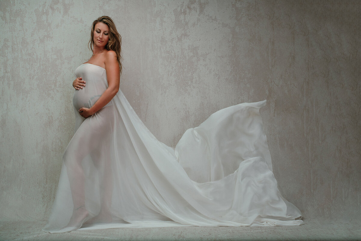 Maternity portrait blonde woman standing wearing white chiffon fabric holding baby bump and looking over shoulder on white textured backdrop