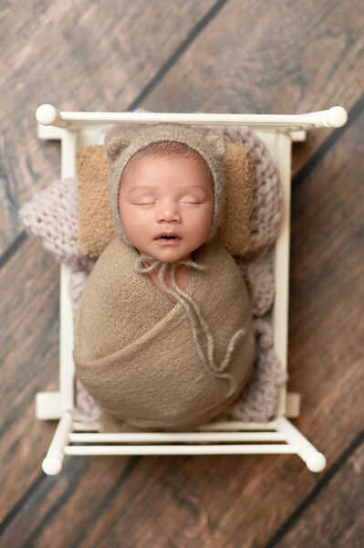 Newborn baby swaddled in cream wrap resting in wooden bowl during Houston newborn photoshoot.