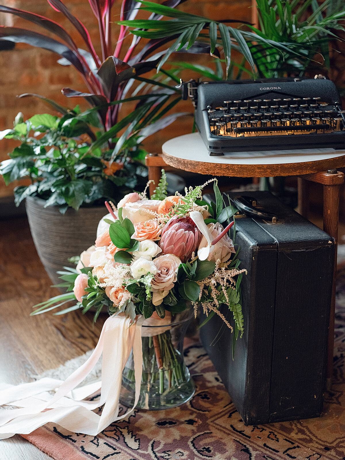 Black antique typewriter on a small midcentury side table next to a tropical bouquet with palm fronds, pink, blush, white and ivory tropical flowers on an oriental carpet with large tropical plants, and palms in the background.