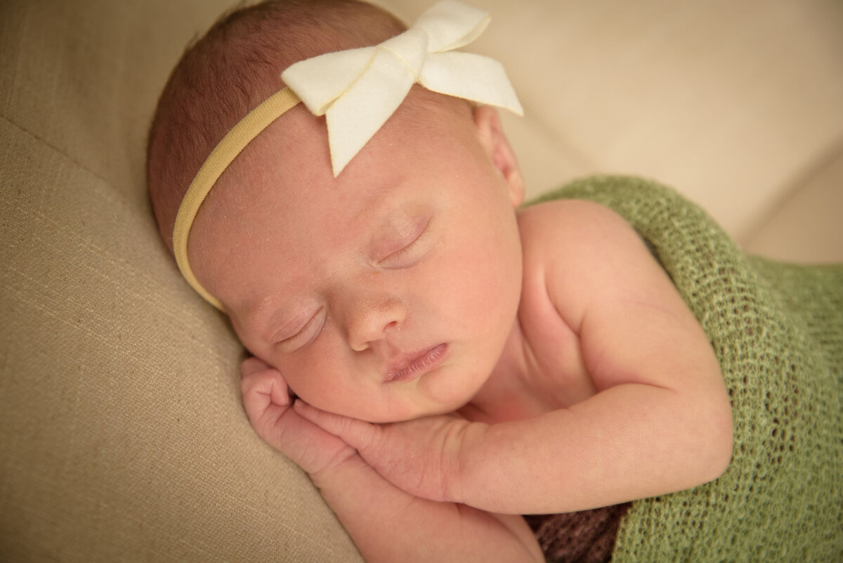 Portrait of newborn baby girl wearing a sage green wrap and cream bow in the nursery of her home in Green Bay, Wisconsin