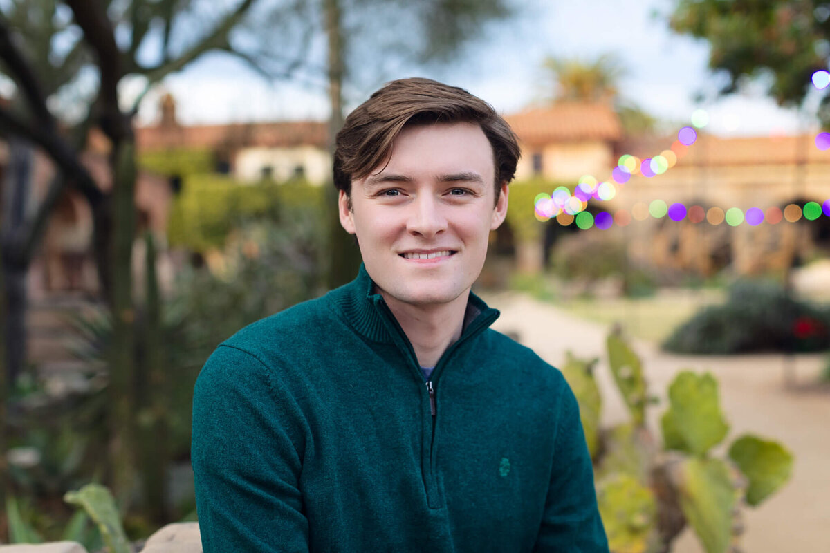 A young man with brown hair, wearing a dark green, long sleeve shirt smiling at the camera in a brick courtyard.