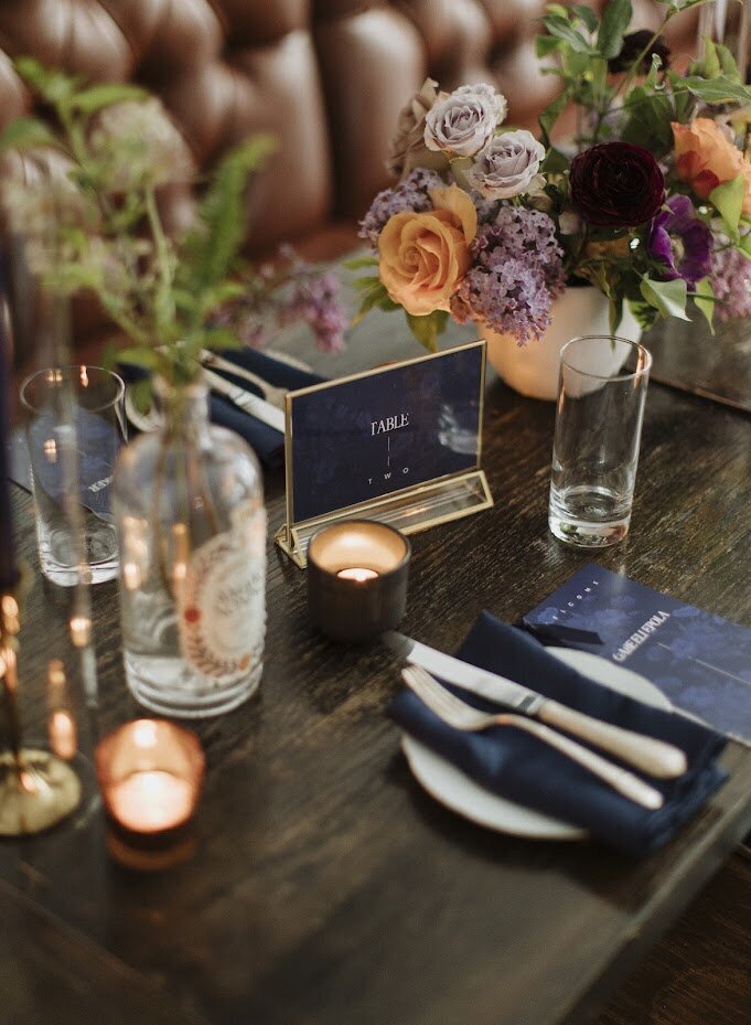 A chic wood table setting featuring a modern navy blue table number, paired with matching navy napkins, soft tea light candles, and a stunning centrepiece of plum, mauve, and mustard-toned florals. This stylish wedding reception design took place at North & Navy in Ottawa, perfectly blending sophisticated details with warm, inviting colors. Ideal for couples seeking modern wedding decor with a personalized touch.