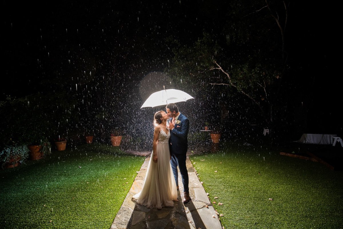 Bride and Groom share a kiss under an umbrella at night with a spotlight on them as the rain comes down