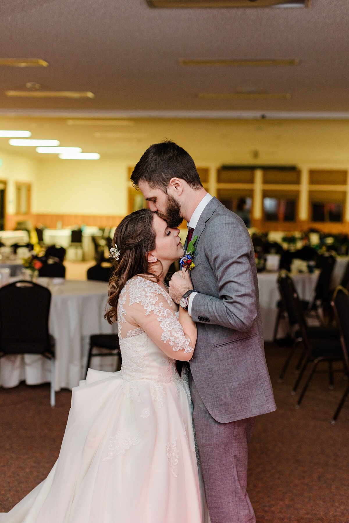 Photo of a bride and groom sharing their private last dance at the end of their wedding reception at the YMCA of the Rockies in Estes Park, Colorado. The bride is on the left and is wearing a long sleeve, intricate, white dress. The groom is on the right and is wearing a grey suit with a boutonniere. As they dance, the groom gives the bride a kiss on her forehead. Empty reception tables can be seen throughout the background.