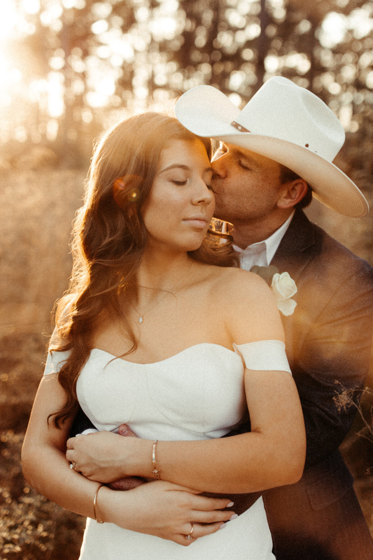 Groom wrapping his arms around his bride and kissing her on the cheek during sunset in the woods