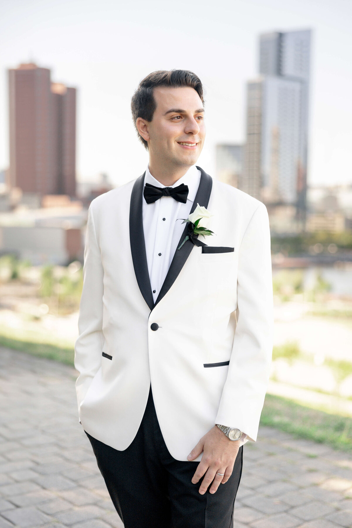 A person wearing a white tuxedo with a black bow tie stands outdoors with a cityscape in the background. The person is smiling and looking to the side.