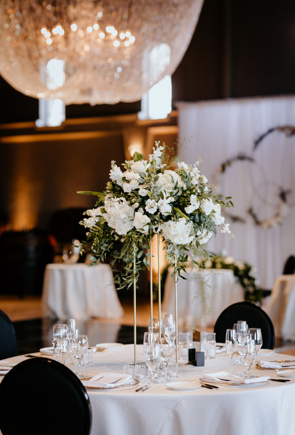 Elegant spring wedding tablescape at the Palm Event Center in Pleasanton, CA, featuring a tall tiered white floral arrangement, white tablecloths, and sleek black chairs.