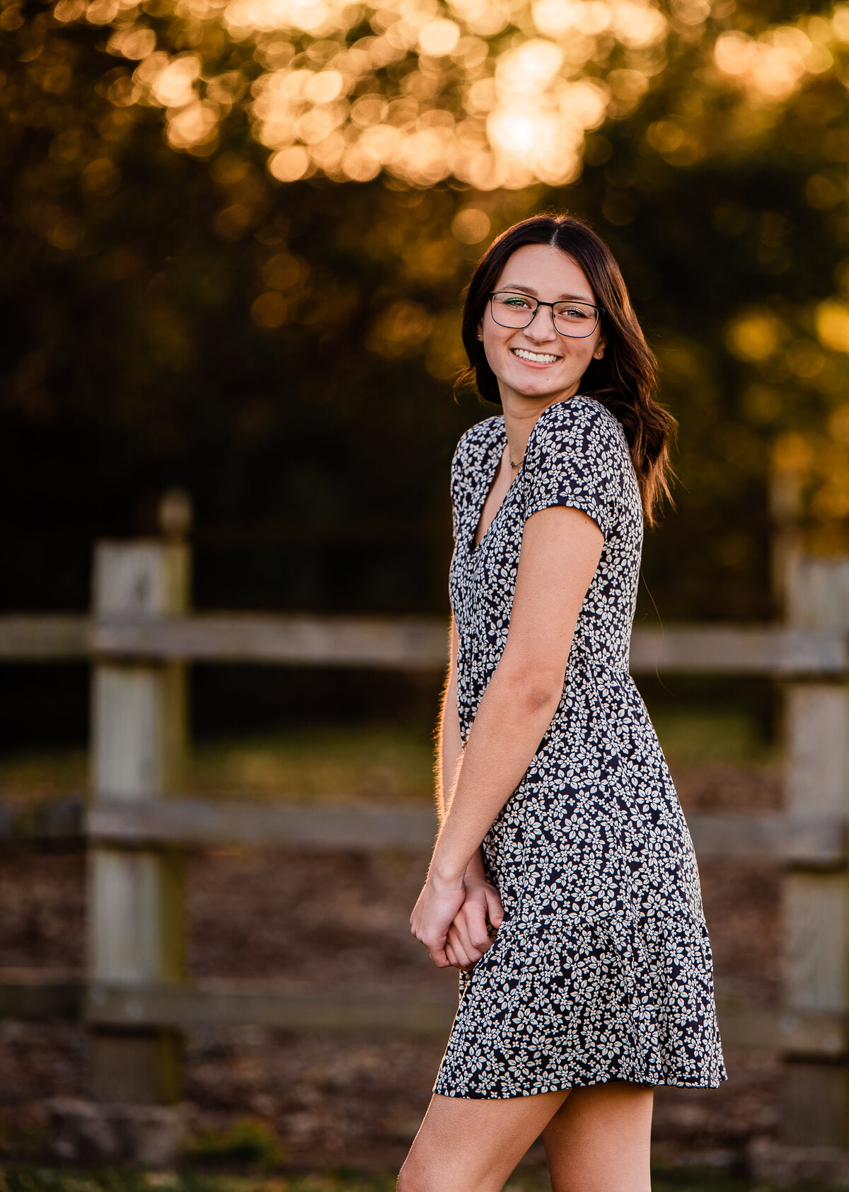A senior in a black floral dress stands in front of a rustic wooden fence at sunset at Ghirardi Park.
